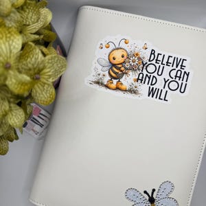 Puede incluir: Una carpeta blanca con una pegatina de una abeja de dibujos animados sosteniendo flores con el texto BELEIVE YOU CAN AND YOU WILL, y un adorno en forma de abeja con alas azul claro brillantes y un cuerpo a rayas negras y doradas.