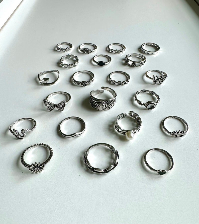 Puede incluir: Un conjunto de 18 anillos de plata con varios dise&ntilde;os, incluyendo mariposas, flores, corazones y patrones geom&eacute;tricos.