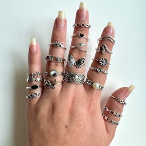 May include: A hand wearing a variety of silver rings with different designs, including a butterfly, a snake, a heart, a flower, a yin yang symbol, and a star. The rings are stacked on multiple fingers.