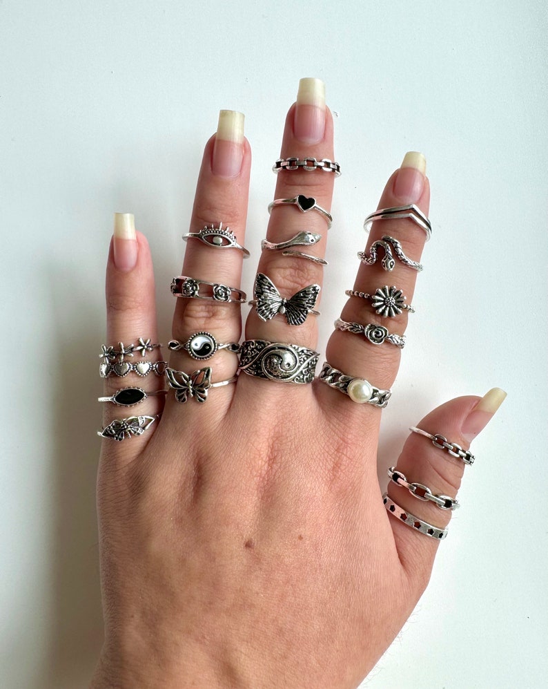 Puede incluir: Una mano con una variedad de anillos de plata con diferentes dise&ntilde;os, incluyendo una mariposa, una serpiente, un coraz&oacute;n, una flor, un s&iacute;mbolo yin yang y una estrella. Los anillos est&aacute;n apilados en varios dedos.
