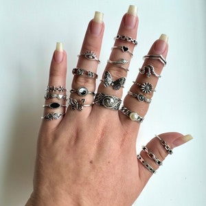 Puede incluir: Una mano con una variedad de anillos de plata, incluyendo una mariposa, una serpiente, un coraz&oacute;n, una flor, un s&iacute;mbolo yin yang y una perla. Los anillos est&aacute;n apilados en cada dedo.