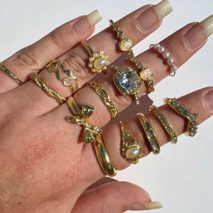 May include: A set of gold-toned rings with various designs, including a bow, a heart, a pearl, and a large clear stone. The rings are worn on a hand with manicured nails.