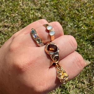 May include: A hand wearing four gold rings. One ring features three oval-shaped, light-colored stones. Another has a large, rectangular amber-colored stone. The third ring has a textured, nugget-like design. The fourth ring has three smaller, oval-shaped, light-colored stones.