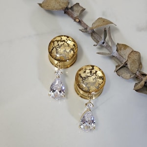 May include: Two gold-coloured ear gauges with a clear top filled with gold leaf flakes. Each gauge has a teardrop-shaped, clear, sparkling gem dangling from it. The gauges are displayed on a white surface with a dried plant stem.