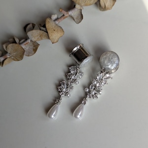 May include: A pair of silver-toned ear gauges with a clear, glittery center and a dangling, ornate design. The design features clear crystals and a pear-shaped pearl drop. A dried plant stem is in the background.