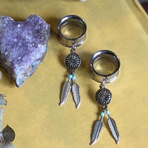 以下が含まれることがあります： 装飾的なデザインが施された銀色の円とターコイズビーズが付いた2本の銀色の羽根がついたチャームがぶら下がっている、銀色の耳ゲージ2個