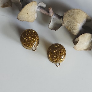 May include: A pair of round gold glitter earrings with small loops. The earrings are set against a white background, with a dried plant stem and leaves.