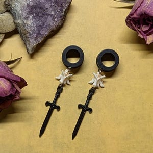 May include: Black gauge earrings with a dangling white bone and a black sword charm. The earrings are set against a tan background with a purple crystal and dried flowers.