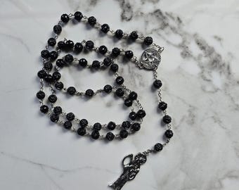 May include: A black rosary with faceted beads and silver-toned metal accents. The rosary features a star-shaped centerpiece and a pendant depicting a figure. The beads are strung on a silver chain.