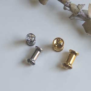 Puede incluir: Cuatro dilatadores de oreja en plata y oro. Dos son plateados con un diseño claro y texturizado, y dos son dorados. Un dilatador dorado tiene un diseño texturizado, y el otro es liso. Los dilatadores están sobre una superficie blanca.