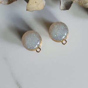 May include: A pair of round stud earrings. Each earring has a gold-coloured border and a small loop. The centre of each earring is filled with white and silver glitter. The background is a light surface with dried leaves.