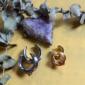 May include: Two pairs of hoop earrings, one silver and one gold. The earrings are on a yellow surface with a purple amethyst crystal.