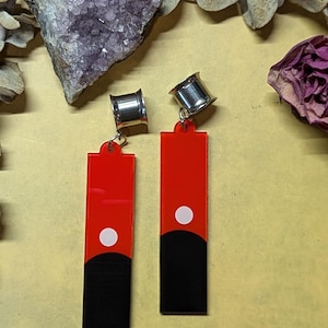 May include: A pair of dangle earrings with a red and black rectangular design and a white dot. The earrings have silver-colored metal plugs. The earrings are displayed on a yellow surface with dried flowers and a purple crystal.