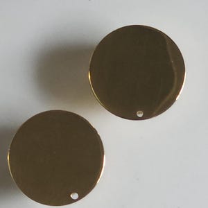 May include: Two shiny, gold-coloured, round metal charms with a small hole near the edge. The charms are reflective and appear to be made of brass or a similar material. The background is a plain, light-coloured surface.