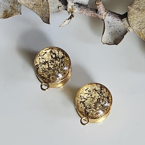 May include: Two gold-coloured, round earrings with clear resin and gold leaf inclusions. Each earring has a small loop at the bottom. A dried branch with leaves is in the background.
