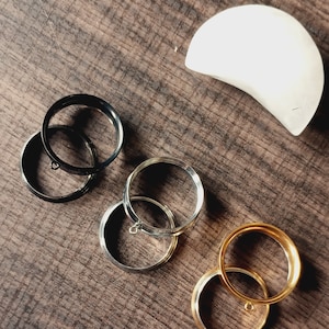 May include: Four metal rings with a decorative loop on each. The rings are stacked on a wooden surface. The rings are black, silver, and gold.