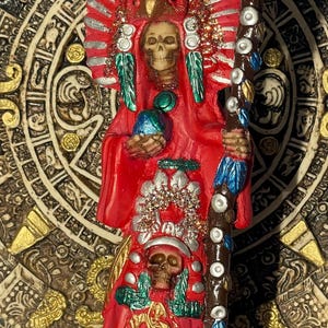 Peut inclure: Statue rouge et argentée d'une figure de squelette en robe rouge, tenant un bâton. La figure porte une coiffe avec des plumes et un visage de crâne. La statue est sur un fond décoratif.