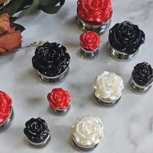 May include: A collection of ear plugs featuring rose-shaped fronts in red, black, and white. Each piece has a metallic base and a sculpted floral design. The set includes various sizes of these decorative plugs arranged in a scattered pattern.