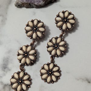 May include: A pair of dangle earrings featuring a floral design. Each earring has three flower-shaped clusters of off-white oval stones set in a bronze-colored metal. The earrings are displayed on a marble surface.