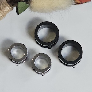 May include: A set of four circular ear tunnels, including two black tunnels and two silver-toned tunnels. Each tunnel features a smooth, cylindrical body with a small, thin metal loop attached to the bottom edge for hanging additional jewelry components.