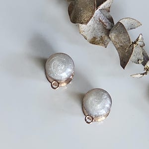 May include: Two small, round earrings with a clear, glittery interior and a gold-coloured base. The earrings have a small loop at the bottom. Dried leaves are in the background.