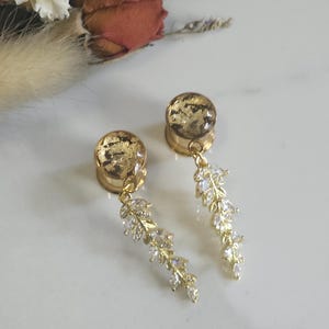 May include: A pair of gold-toned ear plugs featuring a circular face with a mottled gold-leaf effect. Each plug has a dangling gold-colored chain attachment adorned with a series of clear, marquise-cut crystals arranged in a delicate leaf-like pattern.