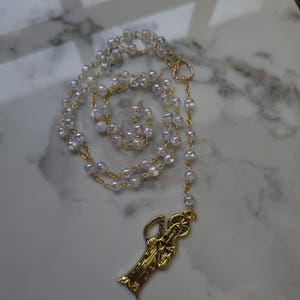May include: A rosary with iridescent, clear beads and gold-colored chain links. The rosary features a gold-colored pendant depicting a religious figure. The beads are strung together in a circular pattern.
