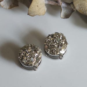 May include: Two silver-coloured, round earrings with a textured, crystal-like surface. The earrings have a dark metal base and a small loop at the bottom. The background is a light, neutral colour with a few dried leaves.