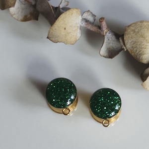 May include: Two round stud earrings with green glitter and gold-coloured metal settings. Each earring has a small loop at the bottom. Dried eucalyptus leaves are in the background.