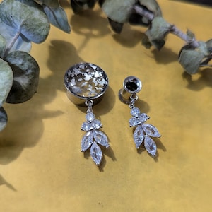 Puede incluir: Un par de dilatadores de oreja de plata con detalles colgantes de cristal. Los dilatadores son redondos y tienen un acabado plateado. Los cristales son transparentes y brillan.