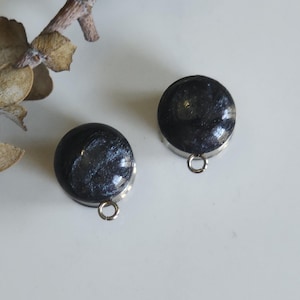 May include: Two round, dark grey earrings with a speckled, glittery appearance. Each earring has a silver-coloured metal base and a small loop. The earrings are displayed on a white surface, with a dried plant stem in the background.