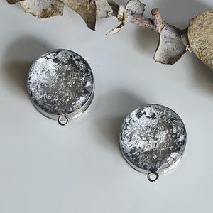 May include: Two circular silver pendants with a clear, glass-like top filled with silver leaf flakes. Each pendant has a small loop at the bottom. The pendants are displayed on a white surface, with a dried plant stem in the background.