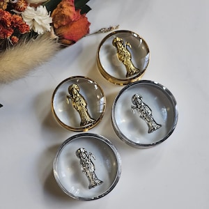 May include: Four circular ear plugs with clear resin centers, each containing a small metallic figure of a robed person holding a scythe. Two plugs feature gold-toned rims and gold-toned figures, while two feature silver-toned rims and figures.
