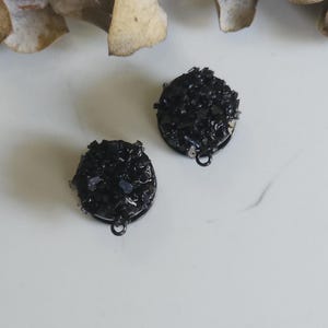 May include: A pair of black druzy stone earrings in a dark metal setting. Each round earring has a small loop at the base. The earrings are displayed on a white surface, with a sprig of dried leaves in the background.