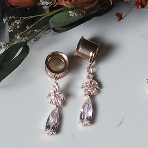 May include: A pair of rose gold ear tunnels featuring dangling charms. Each charm consists of a small cluster of marquise-cut clear crystals attached to a larger pear-shaped teardrop crystal. The metal tunnels have a smooth, polished finish.