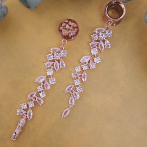 Könnte beinhalten: Ein Paar Ohrstecker aus roségold mit herabhängenden Kristallblatt-Designs. Die Stecker sind mit klaren Strasssteinen verziert und haben eine goldfarbene Oberfläche.