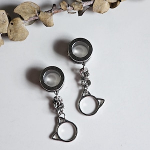 Peut inclure: Deux écarteurs d'oreille argentés avec un charm en forme de chat suspendu à une chaîne. Les écarteurs sont circulaires avec un centre creux. Le charm chat est un contour simple avec un centre circulaire. Le fond est blanc avec une brindille de feuilles séchées.