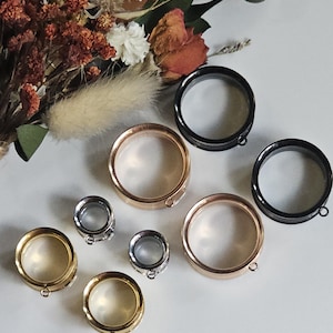 May include: A collection of circular ear gauges in various metallic finishes including gold, rose gold, silver, and black. Each gauge features a hollow center and a small loop attachment at the bottom edge, arranged in a cluster of different sizes.