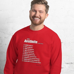 May include: Red sweatshirt with white text that reads "100% Austinite" and a list of things that Austinite's do, including "Born in Austin", "Eat", "Hike", "Help", "Watch", "Drink", "Support", "Survive", "Swim", and "Complain".