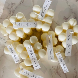 Può includere: Diverse candele bianche a forma di bolla con etichette "Artisan Candle Lab" attaccate. Le candele sono disposte su una superficie di marmo bianco.