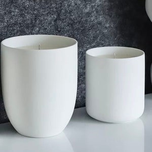 May include: Two white ceramic candles, one taller than the other, with a white wax fill and wicks. The candles are on a white surface.