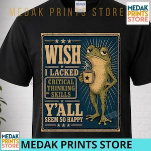 May include: A black short-sleeve t-shirt featuring a graphic print of a green frog holding a coffee mug. The text reads: WISH I LACKED CRITICAL THINKING SKILLS Y'ALL SEEM SO HAPPY. The design is set against a dark blue rectangular background.