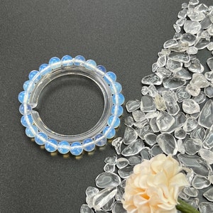 Pode incluir: Um bracelete de contas de opala azul claro com uma banda elástica transparente. O bracelete é rodeado por pedras de quartzo transparentes e uma pequena flor rosa pálida.