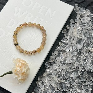 May include: A gold and black gemstone bracelet with round beads. The bracelet is lying on a white notebook with the text "Open Write Dream" printed on it. The notebook is on a black surface with clear quartz chips scattered around it.