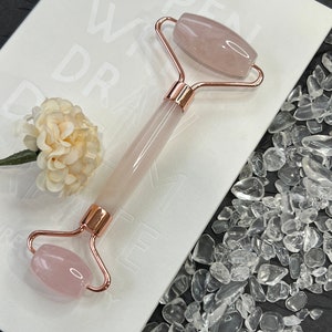 Op de afbeelding: Een rozenkwarts gezichtsroller met een roségouden frame rust op een wit oppervlak met de tekst "OPEN WITH DRA". De roller is omgeven door heldere stenen en een kleine crèmekleurige bloem. De roller is ontworpen voor huidverzorging.