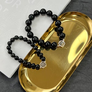 May include: Two black beaded bracelets with silver clasps, one larger and one smaller, on a gold tray. The bracelets are on a white surface with the text "OPEN WRITE DREAM BE" visible.