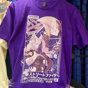 May include: A purple short-sleeve t-shirt featuring a graphic print of characters from Street Fighter. The design includes the text CAPCOM, Japanese characters, and various stylized illustrations of Juri Han, a spider, and a snake within a rectangular frame.