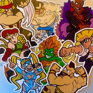 Op de afbeelding: Een verzameling kleurrijke stickers met verschillende personages uit een vechtspel. De stickers tonen personages in dynamische poses, met gedurfde contouren en levendige kleuren. De personages omvatten een verscheidenheid aan vechters met unieke ontwerpen.