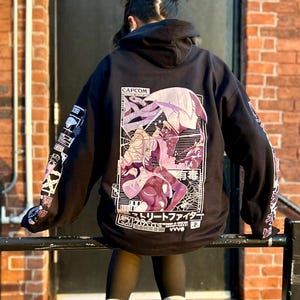 Puede incluir: Sudadera con capucha negra con un estampado gráfico de dos personajes de anime en una pose de lucha. La impresión incluye el texto "CAPCOM" y "ストリートファイター".