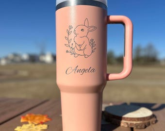 May include: A pink insulated tumbler with a handle and a lid. The tumbler features a laser-etched design of a bunny surrounded by leaves and the name "Angela". The tumbler is sitting on a wooden surface.